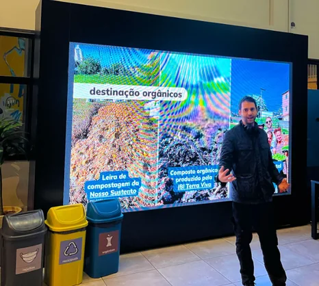 Apresentação sobre destinação de orgânicos diante de um painel, ao lado de contentores de coleta seletiva, na Casa Livre
